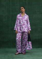 A woman wearing a purple leaf-print silk pajama set with a front pocket and elegant piping around the trouser hems.